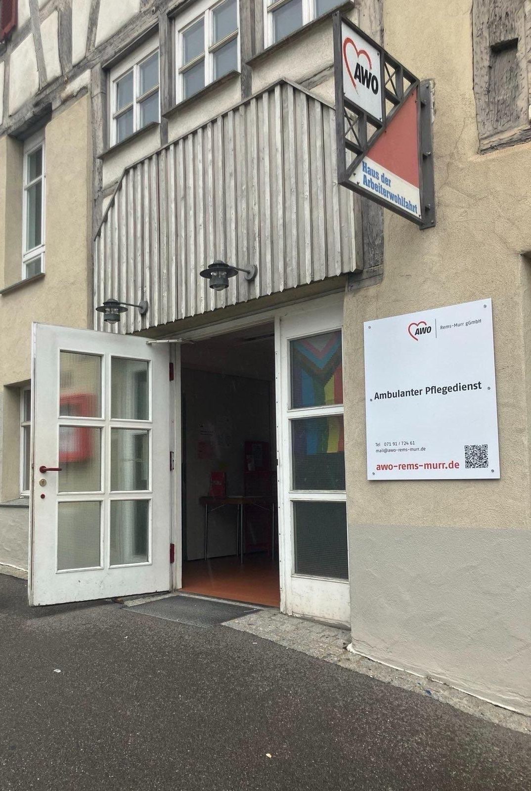 Foto des Eingangs zum AWO Haus Backnang. Das Fachwerkahus hat eine große weiße Tür mit Milchglasscheiben. Die Tür steht offen. Im Fenster rechts neben der Tür hängt eine Regenbogen-Fahne. Daneben hängen an der Hauswand Schilder, die auf das AWO Haus und den ambulanten Pflegedienst hinweisen.