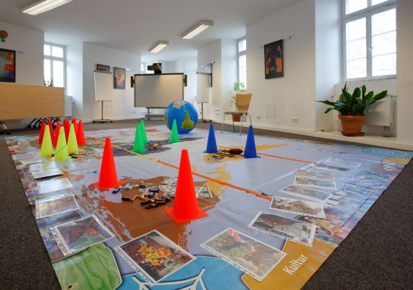 Heller Seminarraum mit digitalem Whiteboard. Auf dem Fußboden ausgebreitete Plakate mit aufgelegten Fotos und Markierungskegel.