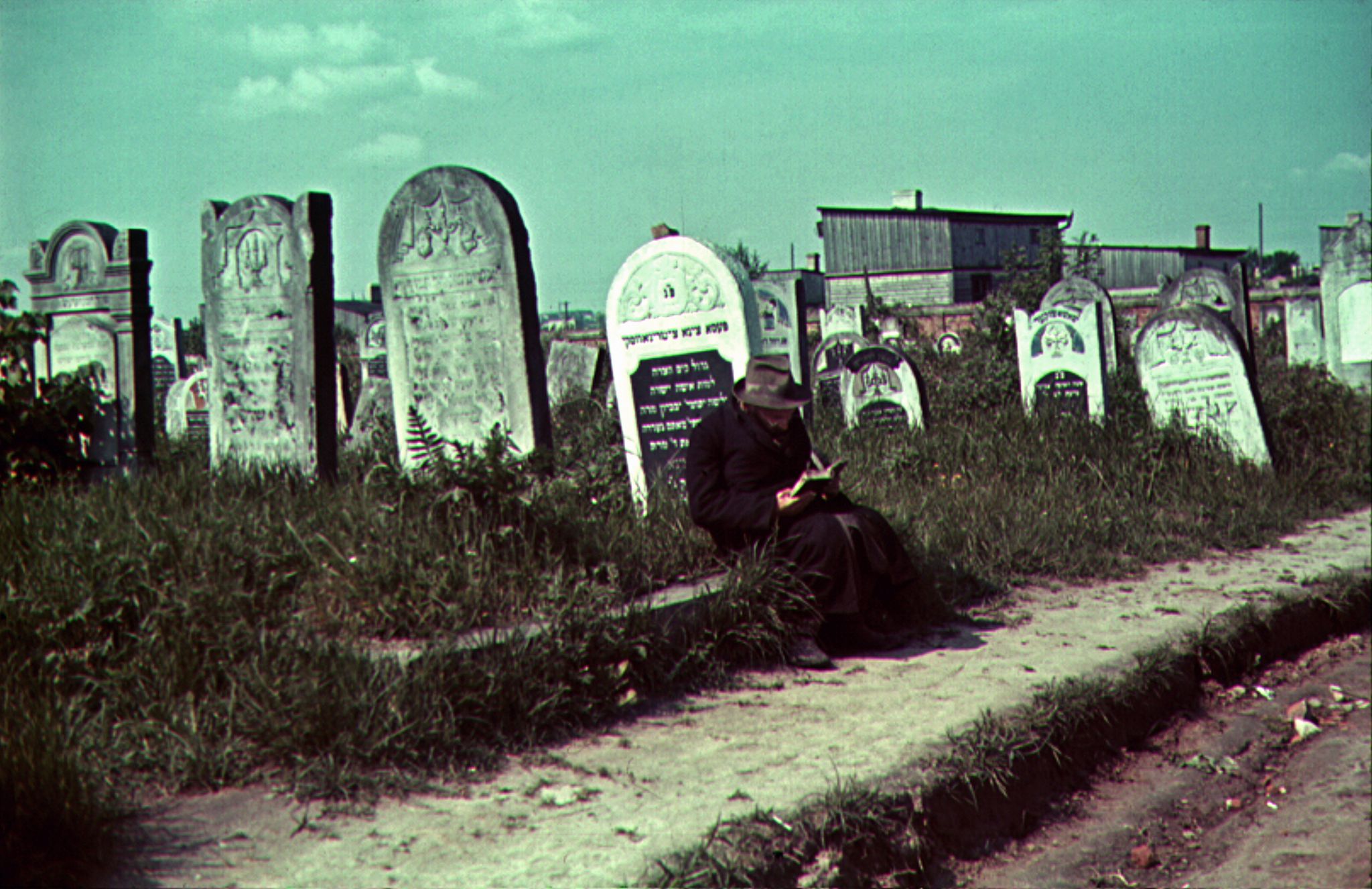 Eine Person sitzt in einem Buch lesend auf einer niedrigen Einfriedungsmauer, hinter ihr stehen alte Grabsteine mit hebräischer Beschriftung