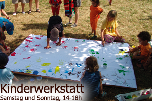Kinder stehen und hocken um ein weißes Leintuch auf der Wiese und drücken mit Fingermalfarben bunte Handabdrücke darauf.