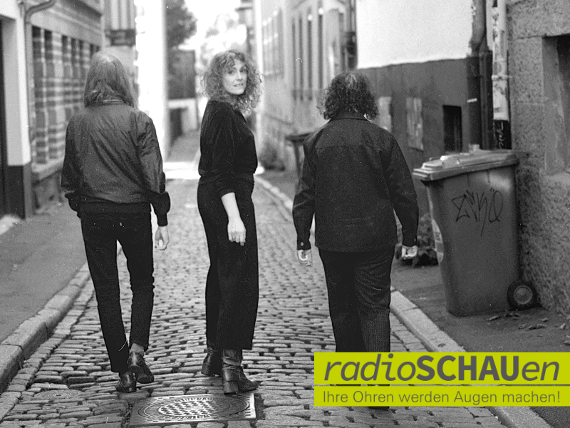 Die Schwarz-Weiß-Aufnahme zeigt drei Personen auf einer Kopfsteinpflastergasse. Zwei Männer gehen mit dem Rücken zur Kamera, während eine Frau in der Mitte lächelnd zurückblickt. Rechts steht ein Müllcontainer mit Graffiti. Unten rechts ist das Logo „radioSCHAUen" zu sehen.