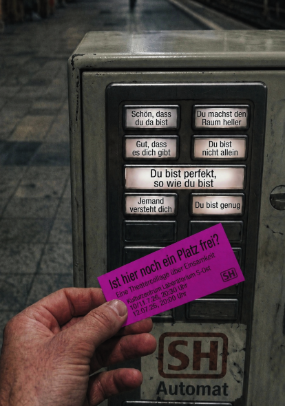 Eine Hand hält ein pinkes Ticket der Theatercollage „Ist hier noch ein Platz frei?“ vor einen Bahnstehautomaten, dessen Tasten durch ermutigende Botschaften wie „Du bist nicht allein“ oder „Gut, dass es dich gibt“ ersetzt wurden. Das Bild verbindet den anonymen öffentlichen Raum mit einer Einladung zu Nähe, Gemeinschaft und Begegnung.