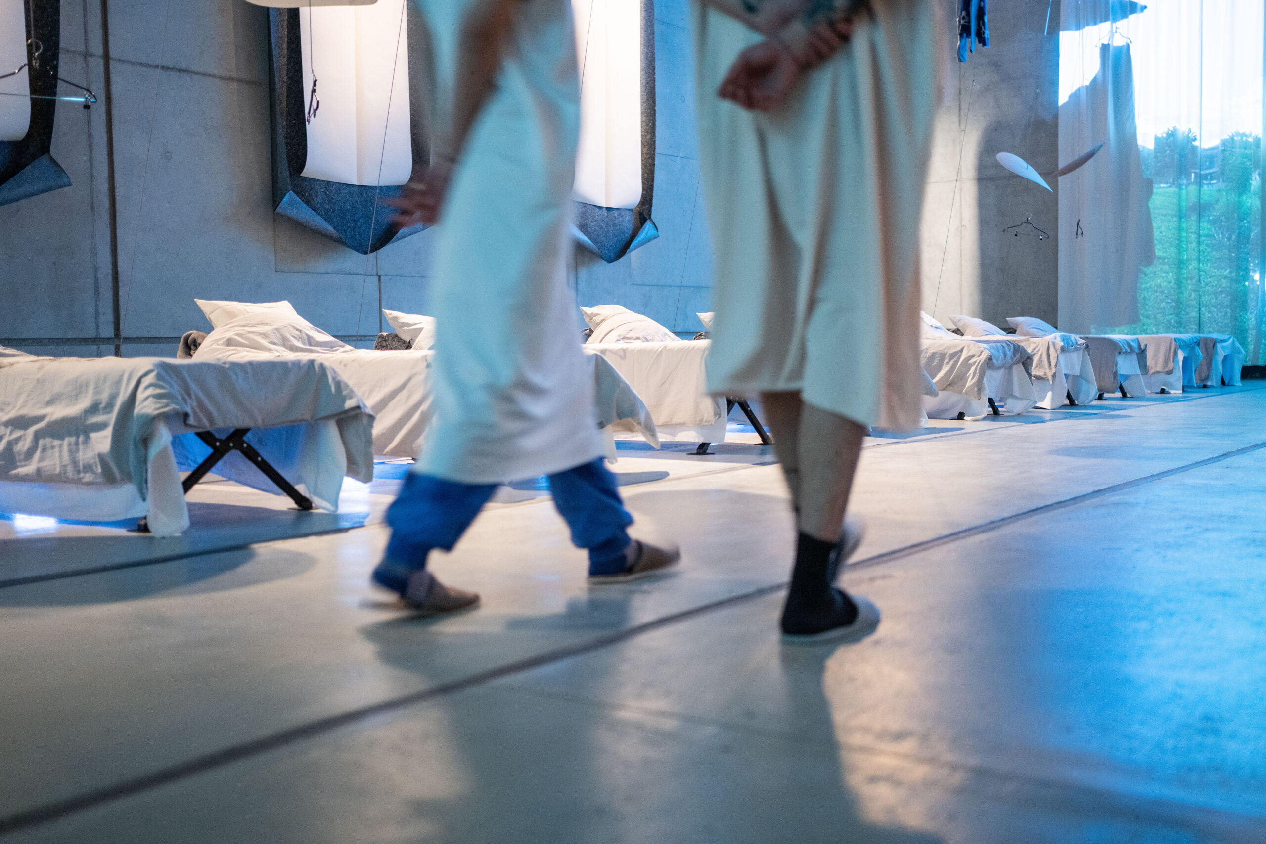 Eine Reihe von Betten steht vor einer Sichtbeton-Wand. Davor laufen zwei Personen von links nach rechts durch das Bild. 